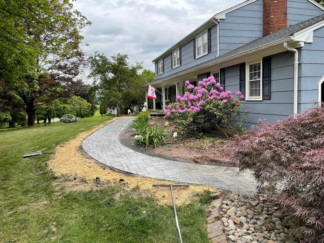 Newly paved stone walkway leading to a blue house with flowering shrubs and lush greenery.