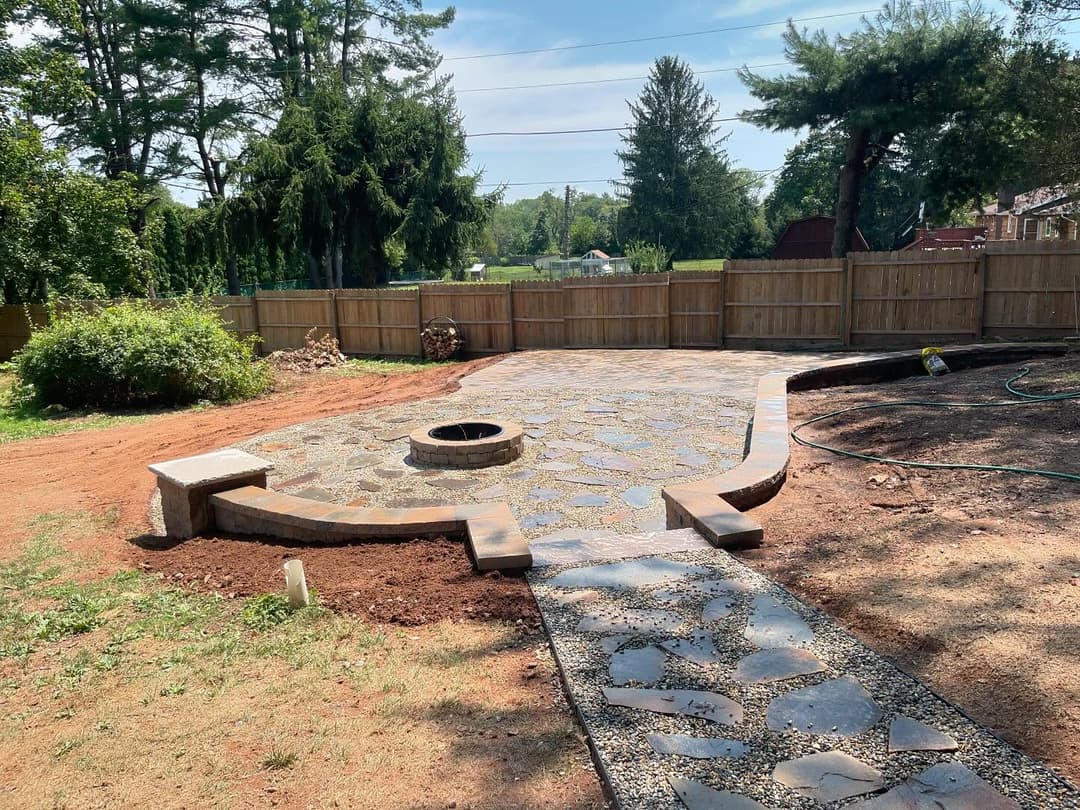 Patio area with stone pathway, fire pit, and fenced backyard in sunny outdoor setting.
