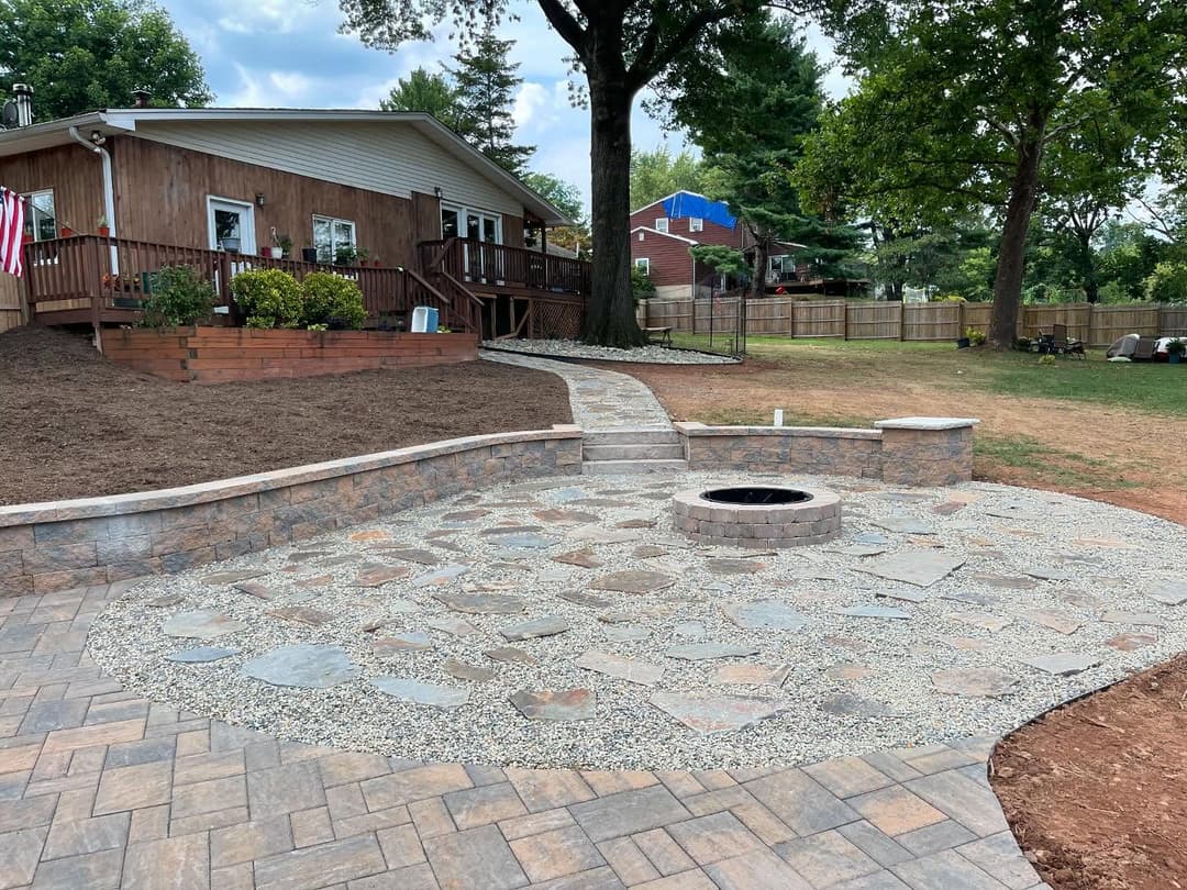 Stone patio with fire pit and winding pathway in backyard with wooden house and lush greenery.