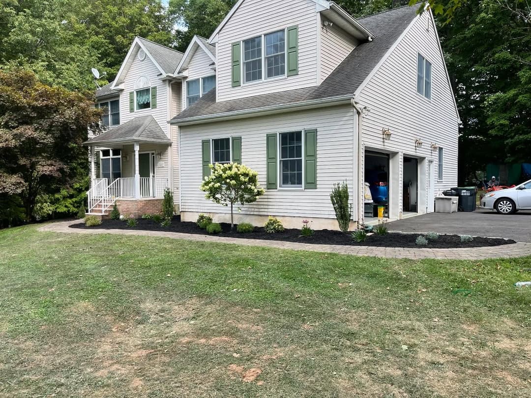Charming two-story house with green shutters, landscaped garden, and driveway on a sunny day.