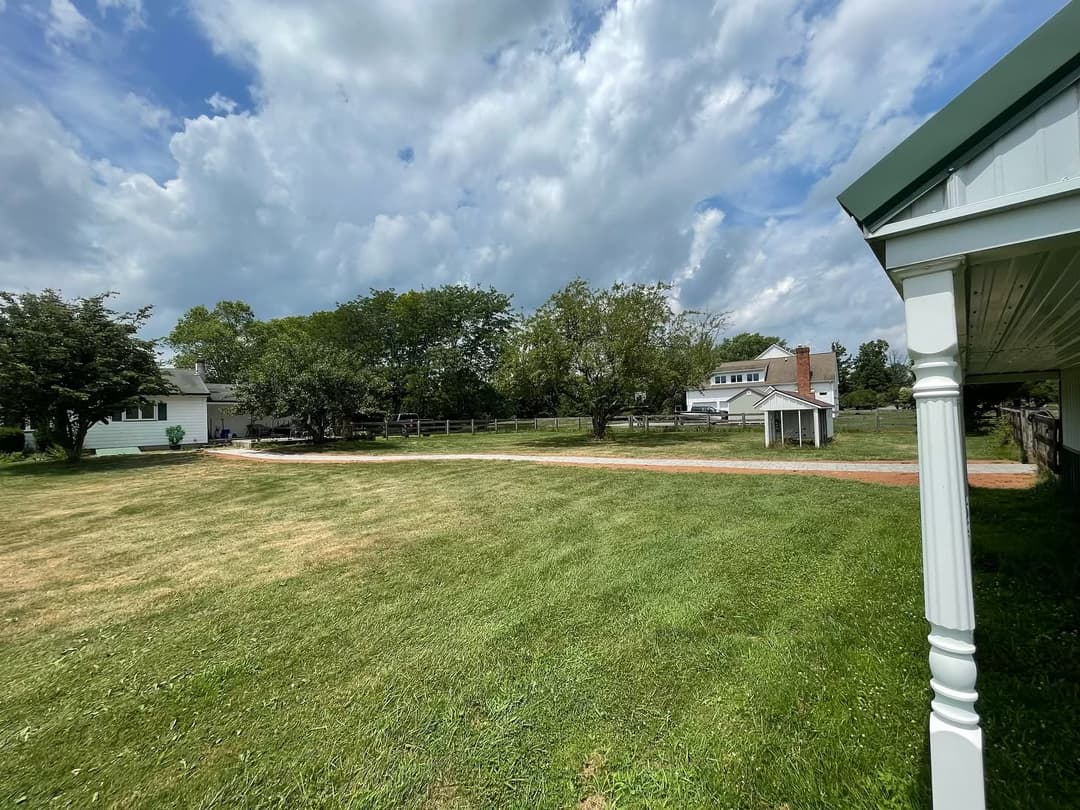 Spacious green yard with a path, trees, and houses under a partly cloudy sky.