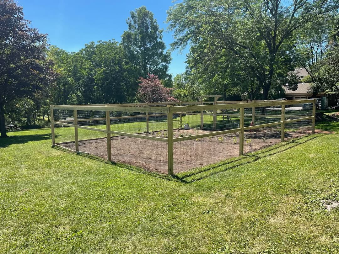 Wooden fence enclosing a garden in a sunny yard, surrounded by trees and greenery.