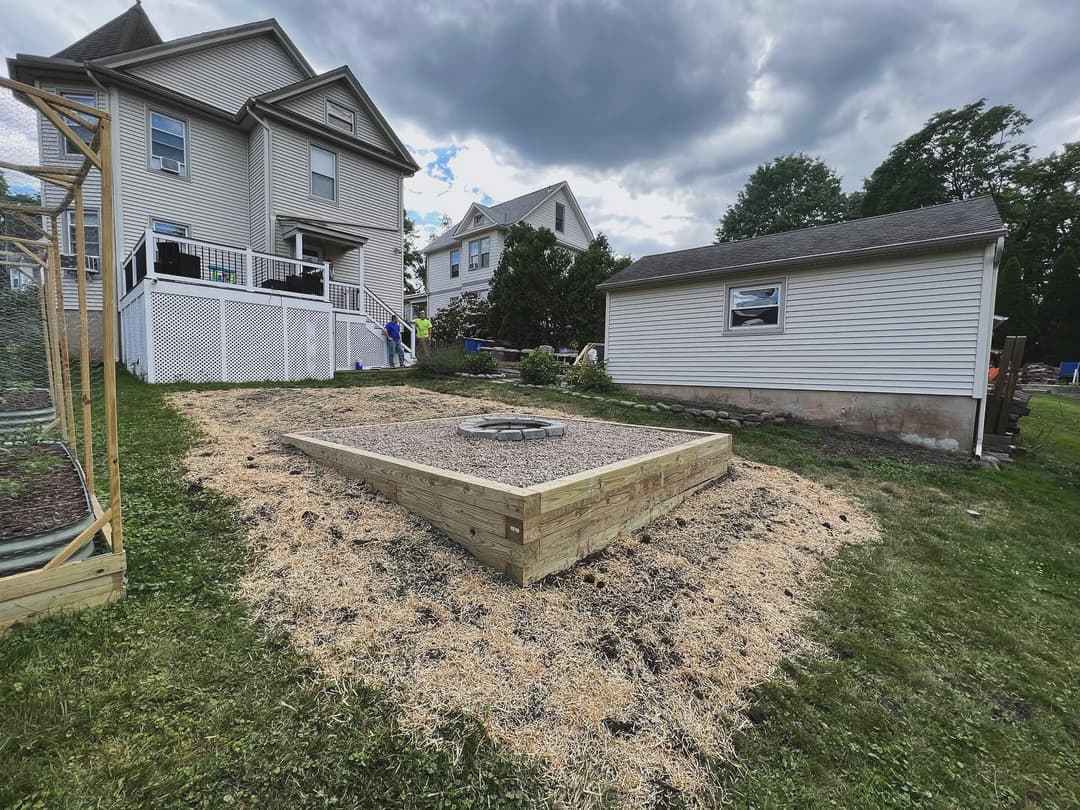 Backyard landscape featuring a wooden platform and a fire pit, with houses in the background.