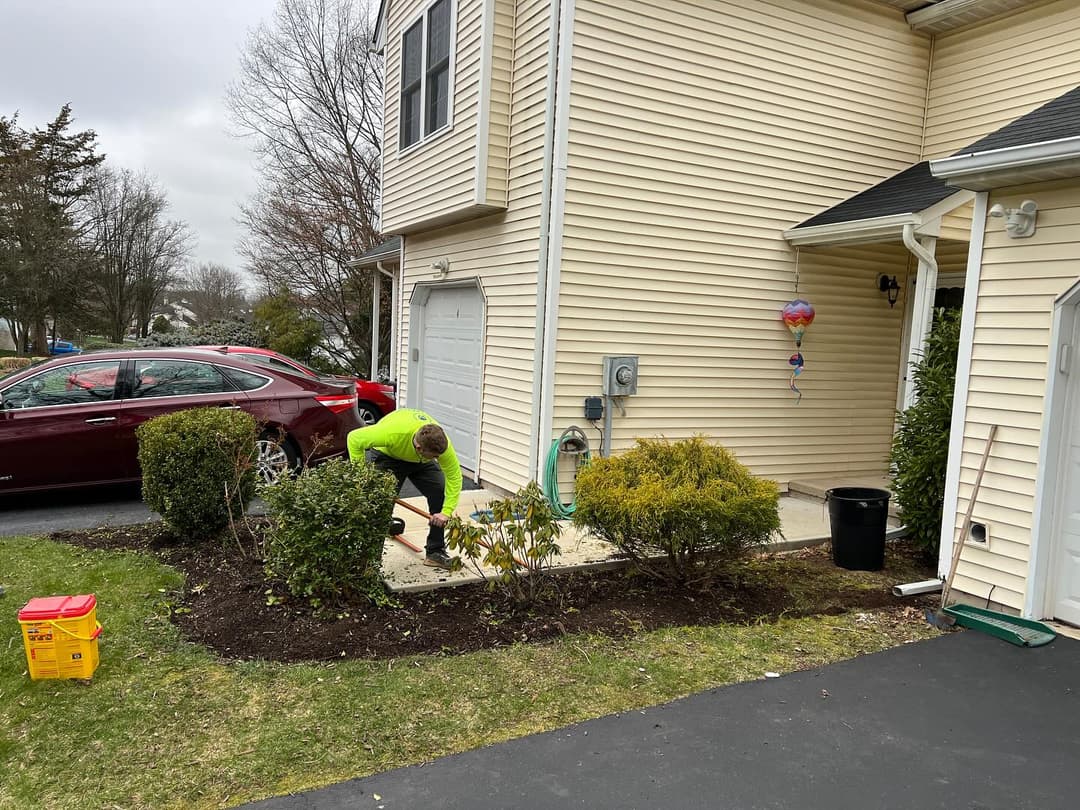 Landscaper trimming bushes near a home's entrance with a car parked nearby.