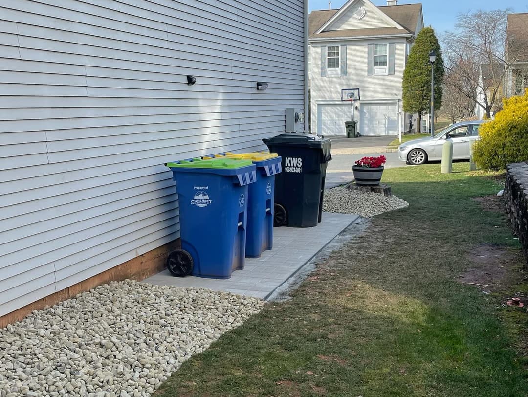 Trash and recycling bins on a paved area beside a house, surrounded by gravel and landscaping.