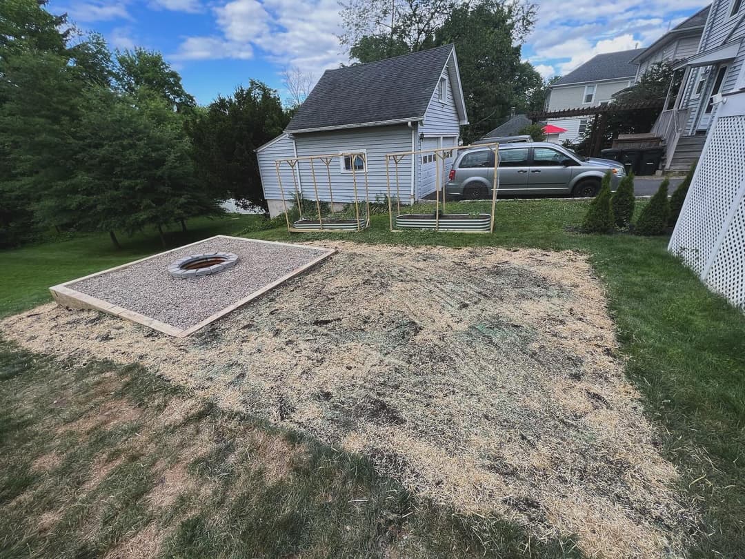 Backyard fire pit area under construction with gravel and wooden border, surrounded by grass.
