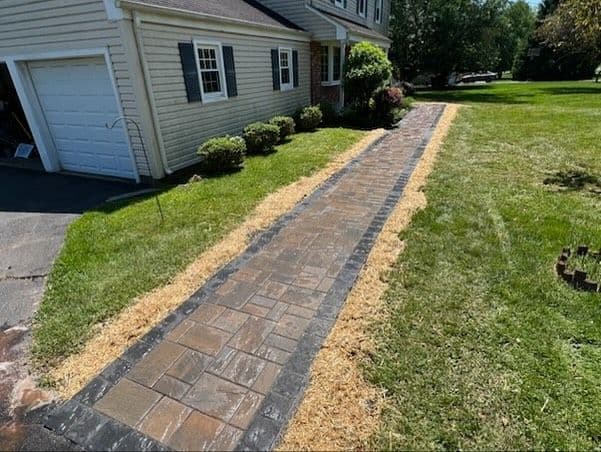 Paved walkway leading to a house, bordered by manicured grass and landscaping.